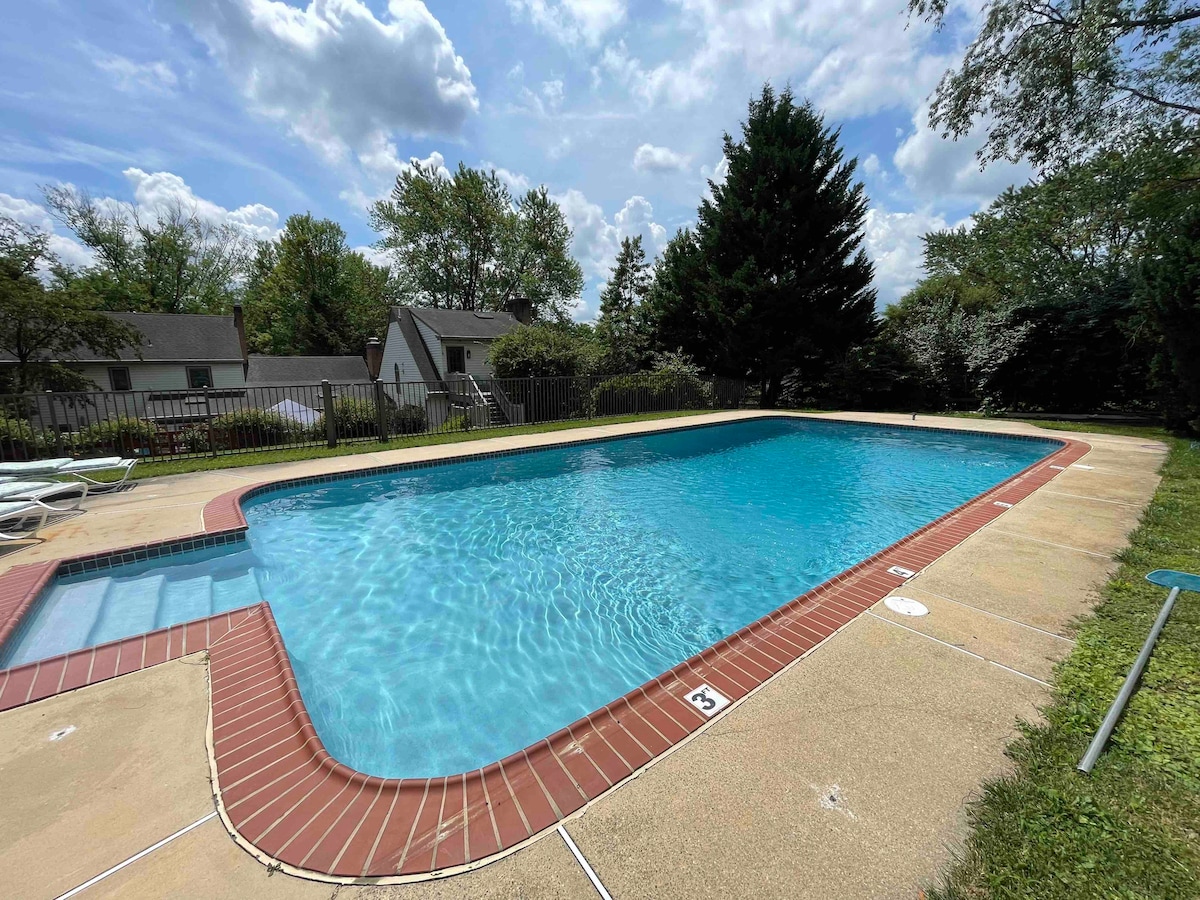 The image displays a large swimming pool set within a well-maintained outdoor area. Sun loungers surround the deck, and greenery serves as a backdrop, adding to the serene atmosphere. The clear blue water reflects the sky, creating a refreshing appearance.