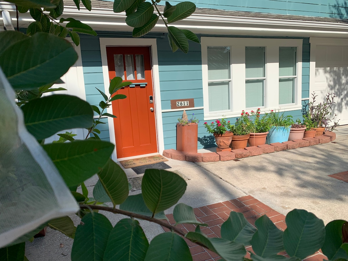 The entrance is highlighted by a vibrant orange door framed by several windows. Flower pots containing blooming plants line the walkway, while a small garden area adds charm to the exterior. Lush green leaves partially frame the scene, suggesting a welcoming atmosphere.