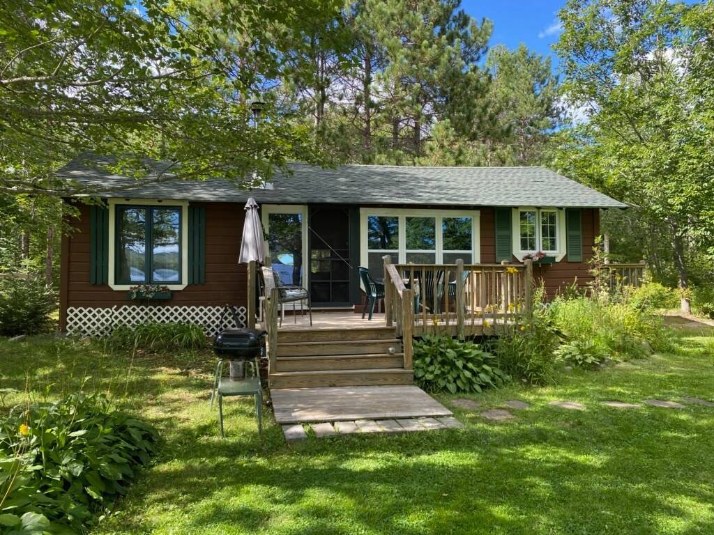 A rustic cottage is surrounded by greenery, featuring a wooden front deck with steps leading up to the entrance. A charcoal grill is positioned nearby, and patio furniture is set for outdoor seating. Large windows provide views of the lake beyond the trees.