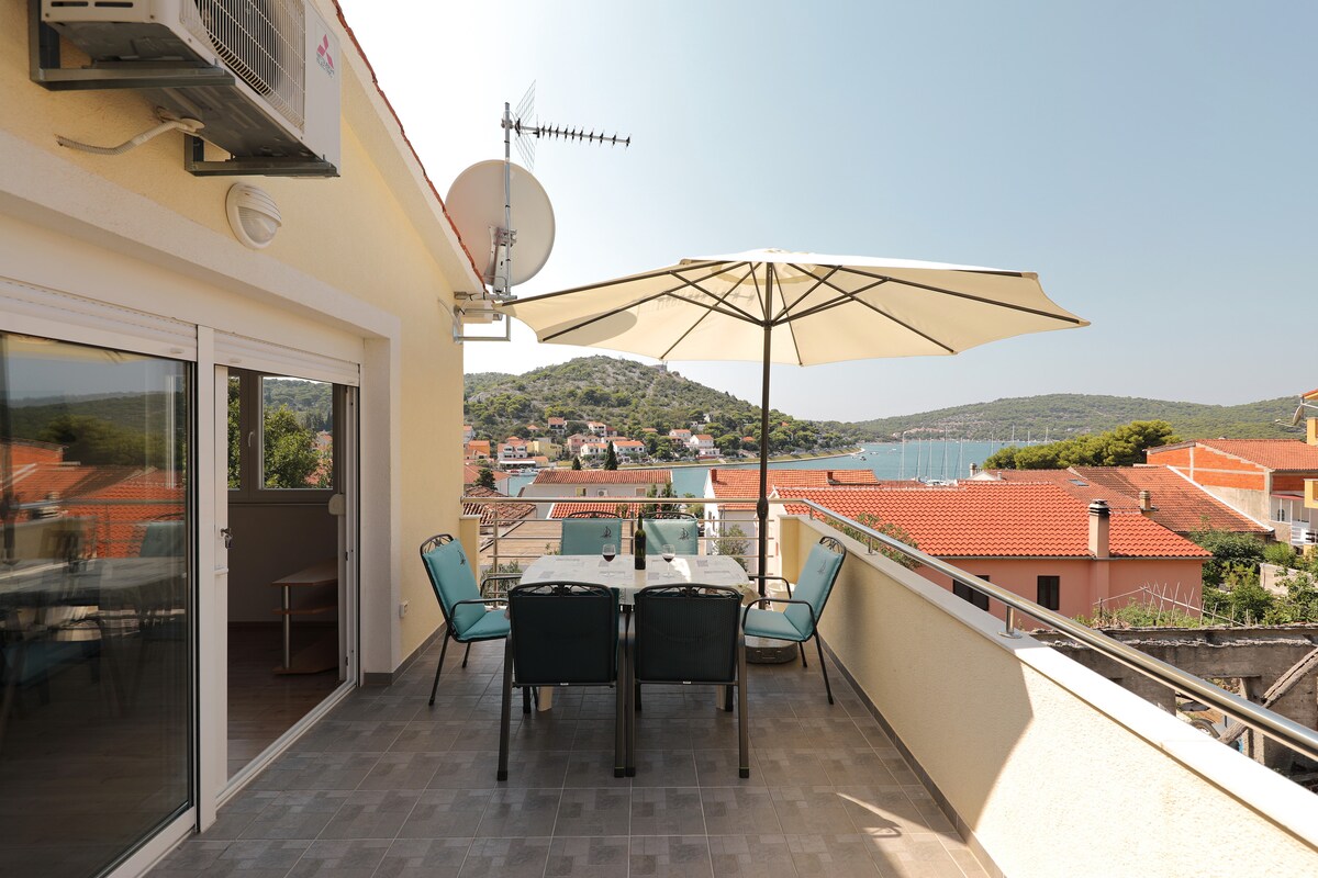 A terrace is shown with a dining table and several chairs. An umbrella provides shade, and a view of the sea and surrounding hills can be seen in the distance. The setting offers a spacious area for relaxation.