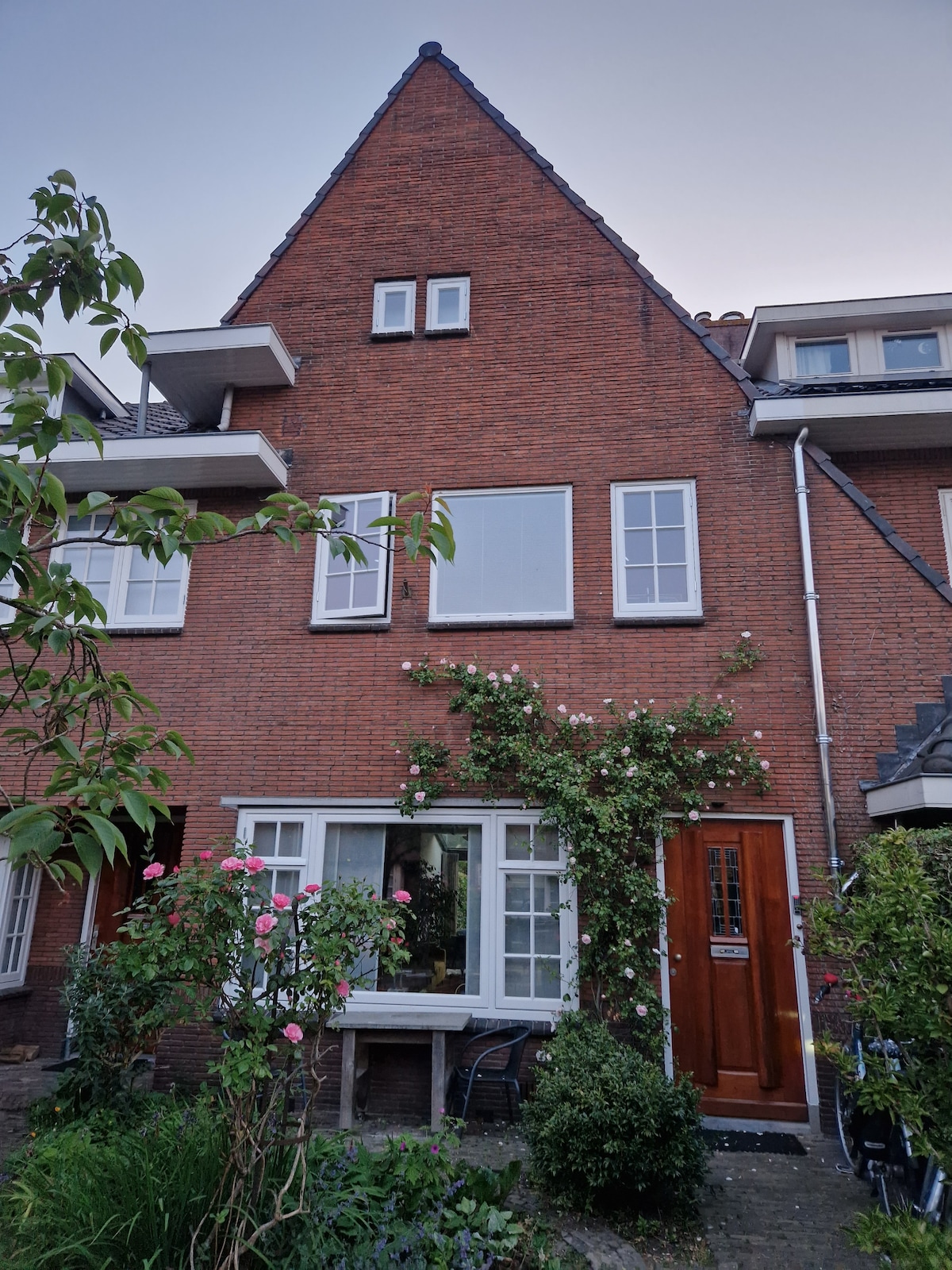 The exterior of a historic brick house is showcased, featuring a peaked roof and large windows. A doorway is framed by lush greenery, with flowering plants adding a touch of color. Two chairs are positioned outside, inviting relaxation in the front garden.