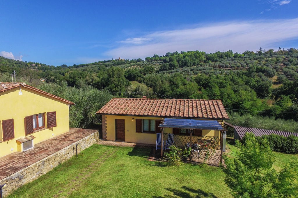 A charming single-story house is set amidst lush greenery, featuring a terracotta roof and a welcoming porch. The surrounding landscape showcases rolling hills and olive trees, contributing to a serene rural environment. A sunlit outdoor space is perfect for relaxation.