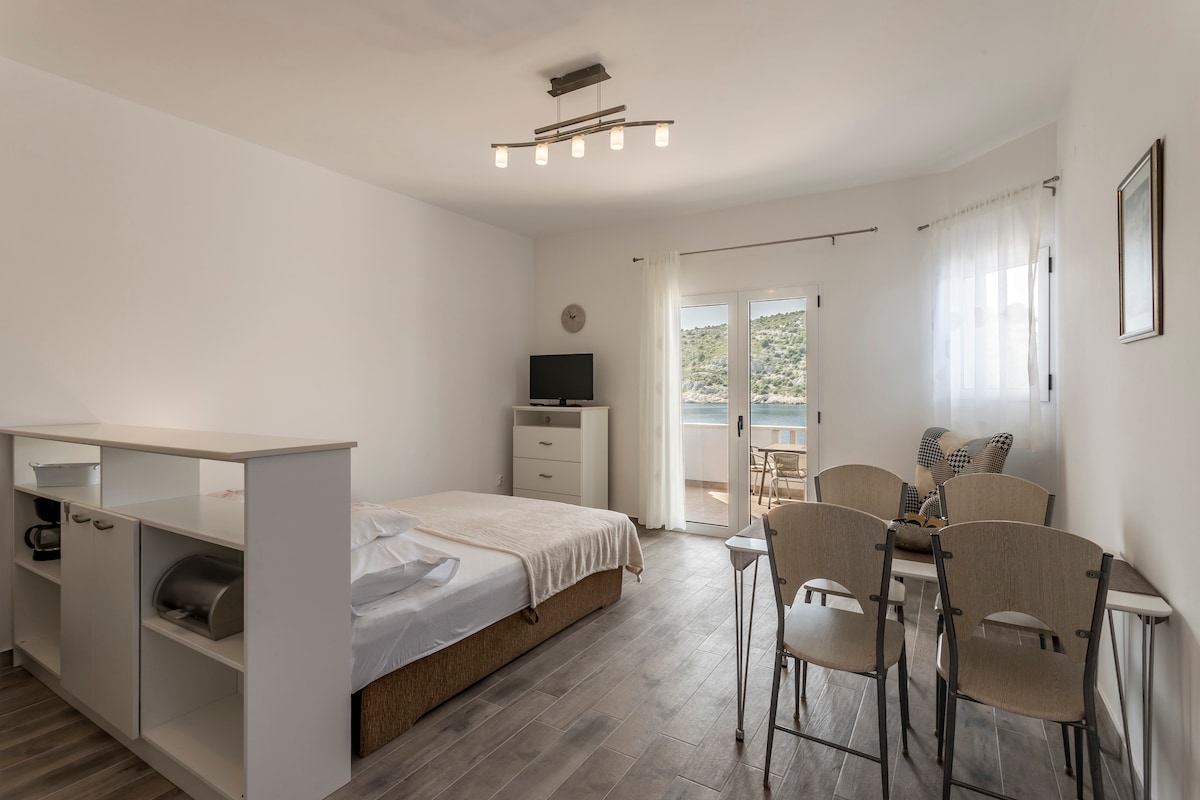A spacious room features a comfortable bed with a light coverlet, accompanied by a small table with two chairs. Natural light filters through a partially open sliding door, revealing a scenic view. A television is positioned near a dresser, providing convenience for guests.