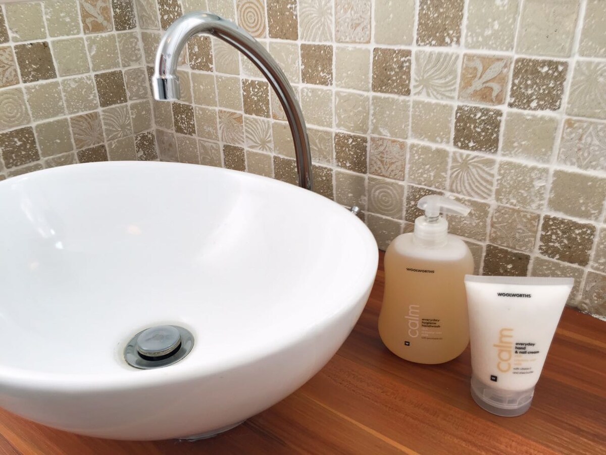 A modern sink made of white ceramic is positioned beside a textured beige tile wall. Two bottles of toiletries are placed on the wooden countertop, one pump bottle and one tube, both designed for personal care.