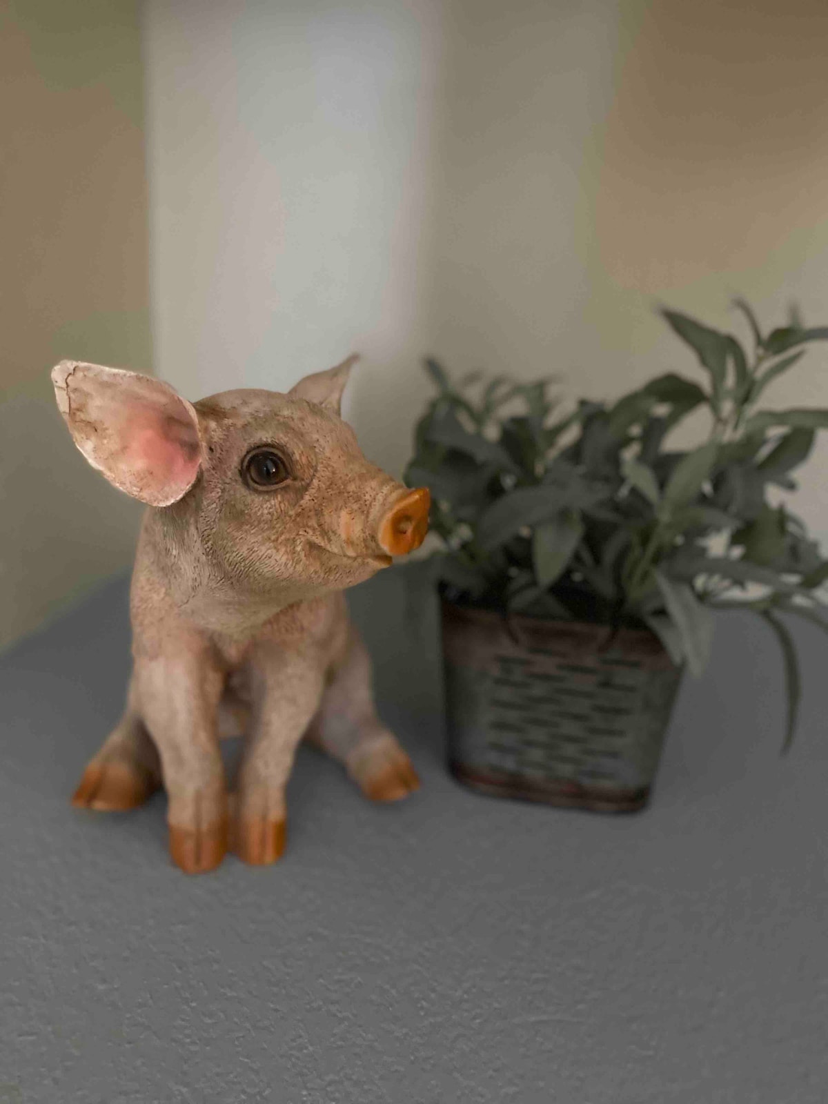 A decorative figurine of a pig sits next to a small potted plant. The pig is positioned with an inquisitive expression and features pink-tinted fur and orange accents on its snout and paws. The plant provides green foliage, complementing the charming, country-themed decor.