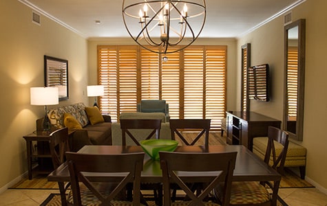A welcoming living area is featured, with wooden furniture including a dining table and chairs. Natural light filters through window shutters, illuminating the space. A comfortable sofa and accent chair offer seating, while a decorative chandelier and wall-mounted television add functionality.