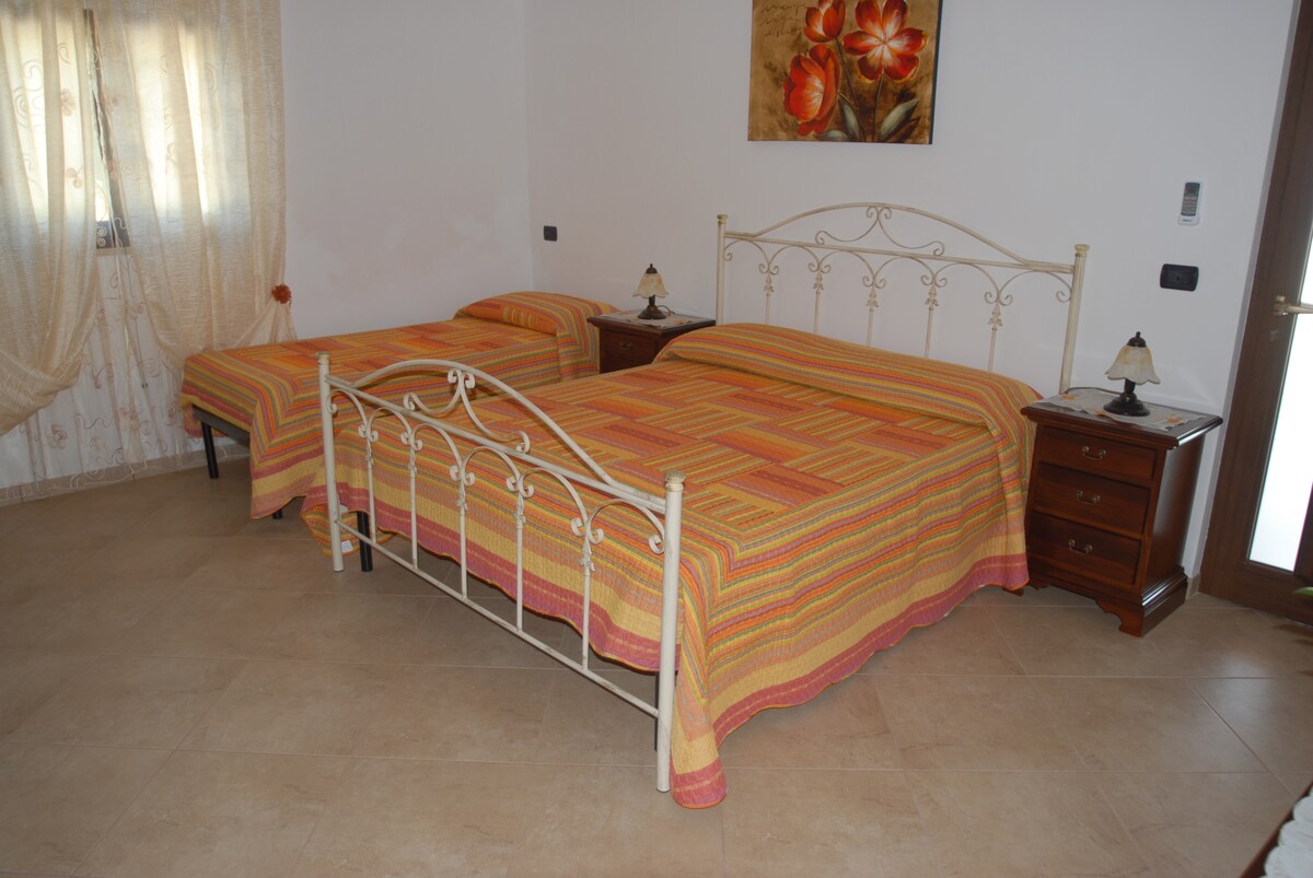 A bright bedroom features a queen-sized bed with a colorful striped blanket and a single bed alongside. Two nightstands with lamps are positioned next to the beds. Soft, natural light filters through sheer curtains, illuminating the neutral-toned floor and wall art.