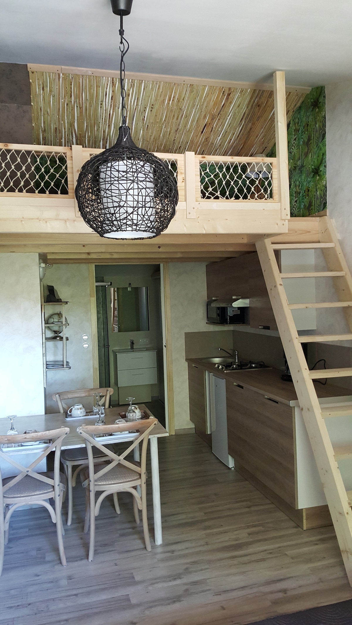 A view of the open living space showcases a dining table set with dishware. A compact kitchen is equipped with essential appliances. A staircase leads to a cozy loft area, while natural elements are incorporated in the decor, creating a warm and inviting environment.
