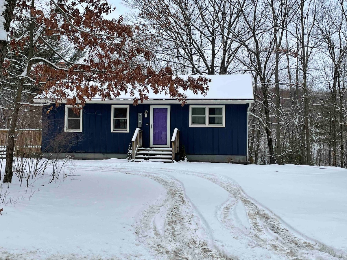 A charming blue cabin is nestled in a snowy landscape, framed by leafless trees. The entrance features a welcoming purple door, and the pathway leads to the front steps, providing a cozy and inviting entryway. Snow covers the ground and accents the surrounding scenery.