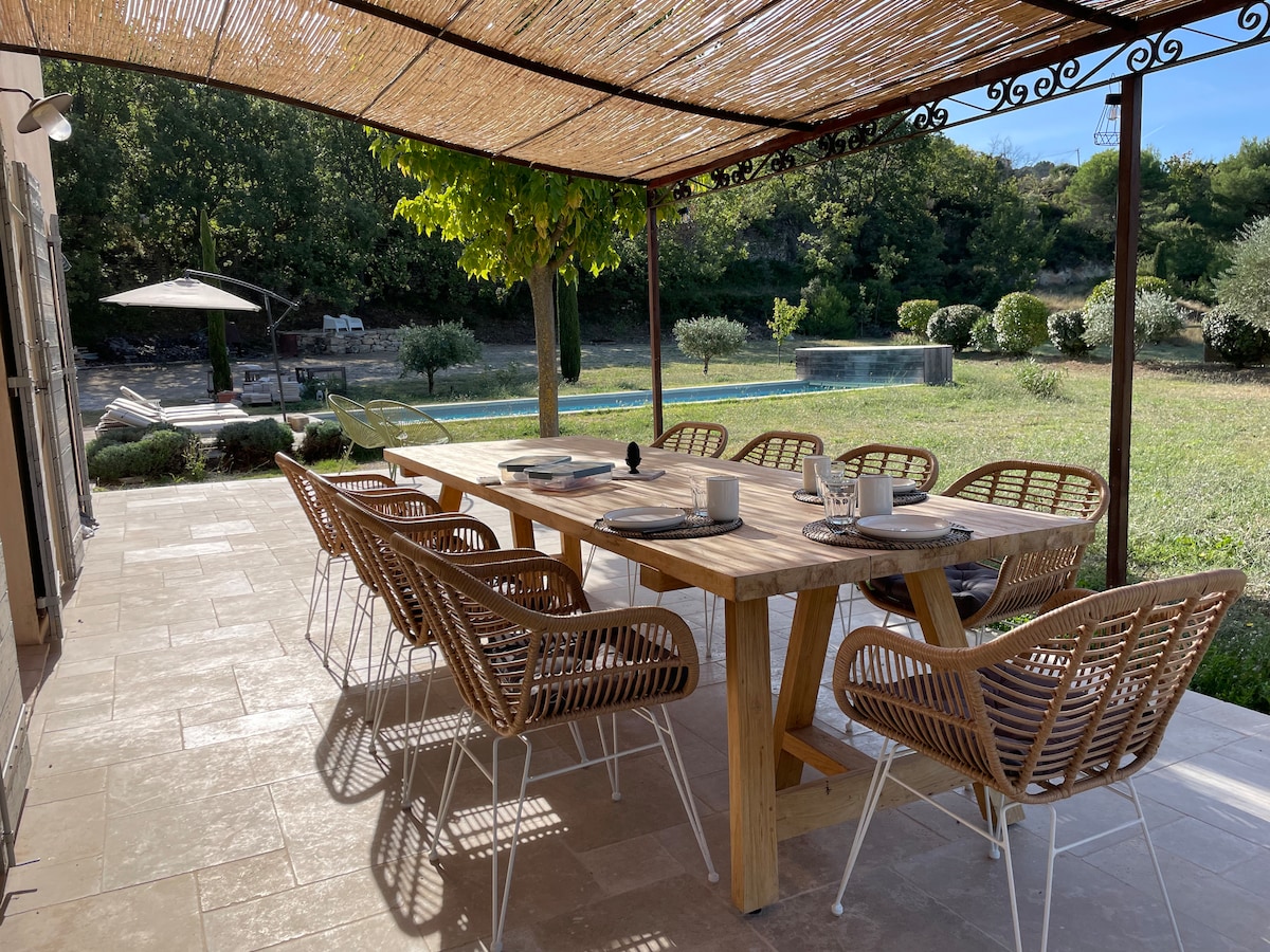 An outdoor dining area features a large wooden table surrounded by eight wicker chairs, shaded by a bamboo covering. A view of the landscaped garden and a swimming pool can be seen in the background, enhancing the inviting space for meals al fresco.