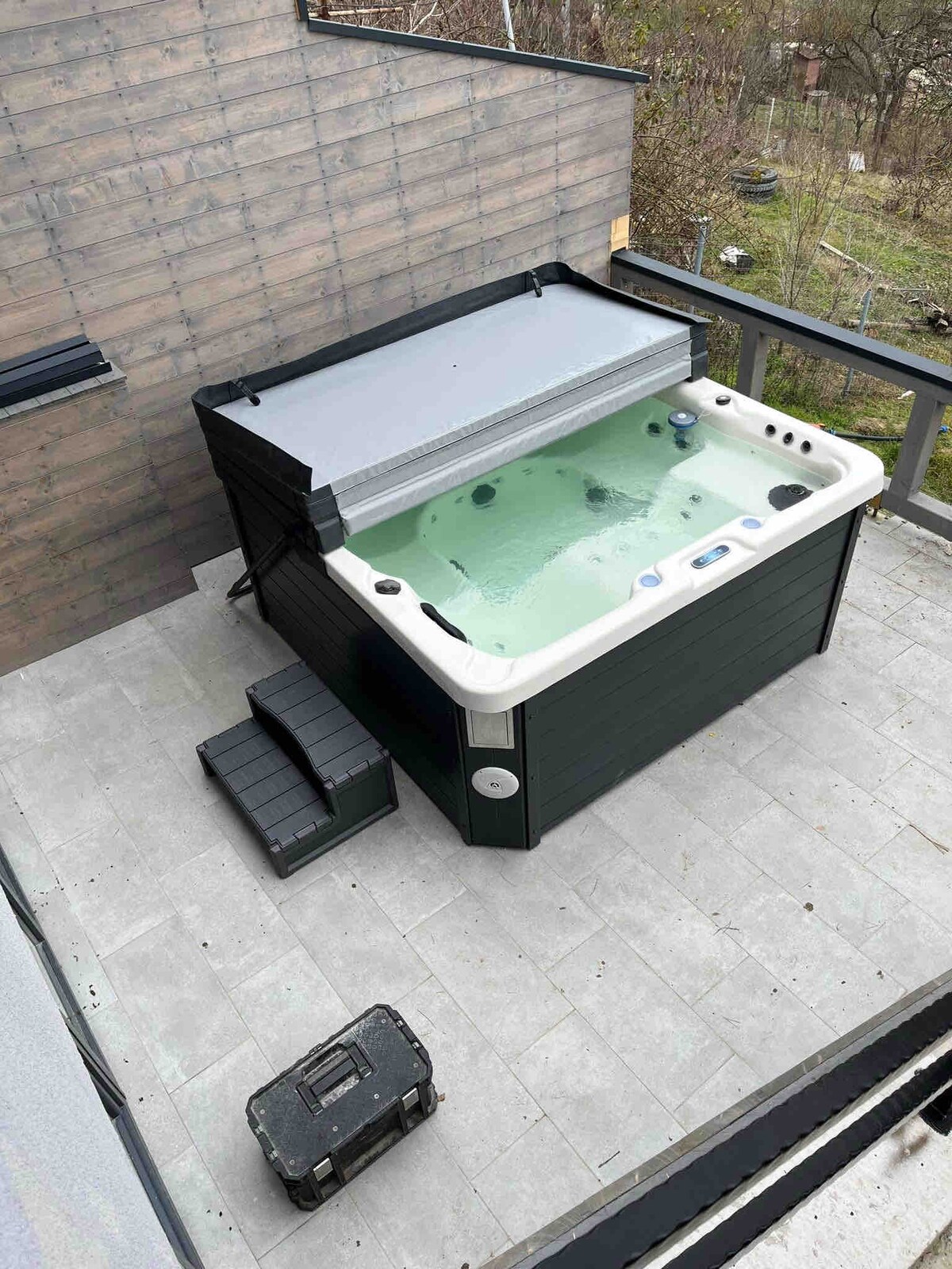A spacious deck is shown featuring a hot tub with a cover. Steps lead up to the tub, surrounded by a stone surface. A small storage box is located nearby, enhancing the outdoor space's functionality.