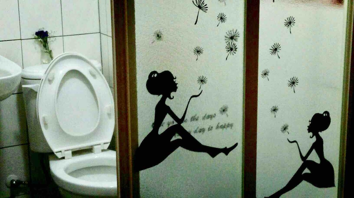The bathroom features a frosted glass partition adorned with artistic silhouettes of a seated figure and a dancer among whimsical dandelion designs. A flower arrangement on the countertop adds a touch of color, complementing the neutral tones of the tiled space.