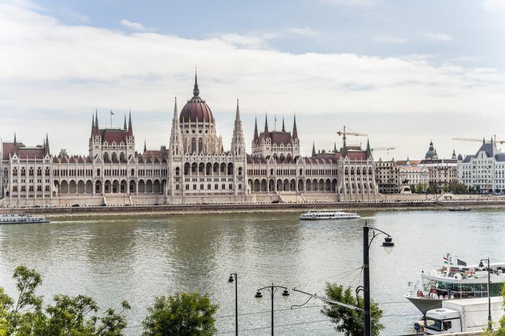 The image features the iconic Hungarian Parliament Building, with its distinctive dome and turrets reflected in the calm waters of the Danube River. Nearby boats are seen along the water, framed by greenery and urban elements in the foreground.