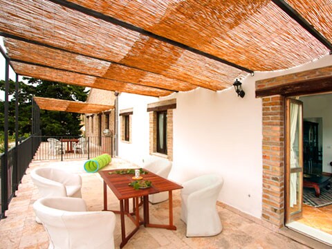 A spacious terrace is covered by a natural overhead canopy, providing shade for a wooden table surrounded by four white armchairs. Large glass doors open to the interior, while the areas beyond the railing showcase greenery and the surrounding landscape.