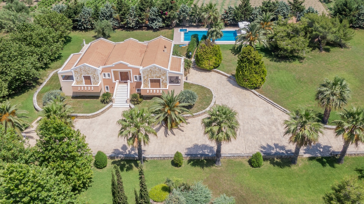An aerial view of a spacious villa surrounded by landscaped gardens featuring numerous palm trees. A private pool can be seen in the background, complemented by a well-maintained driveway leading up to the entrance. The lush greenery enhances the serene environment.