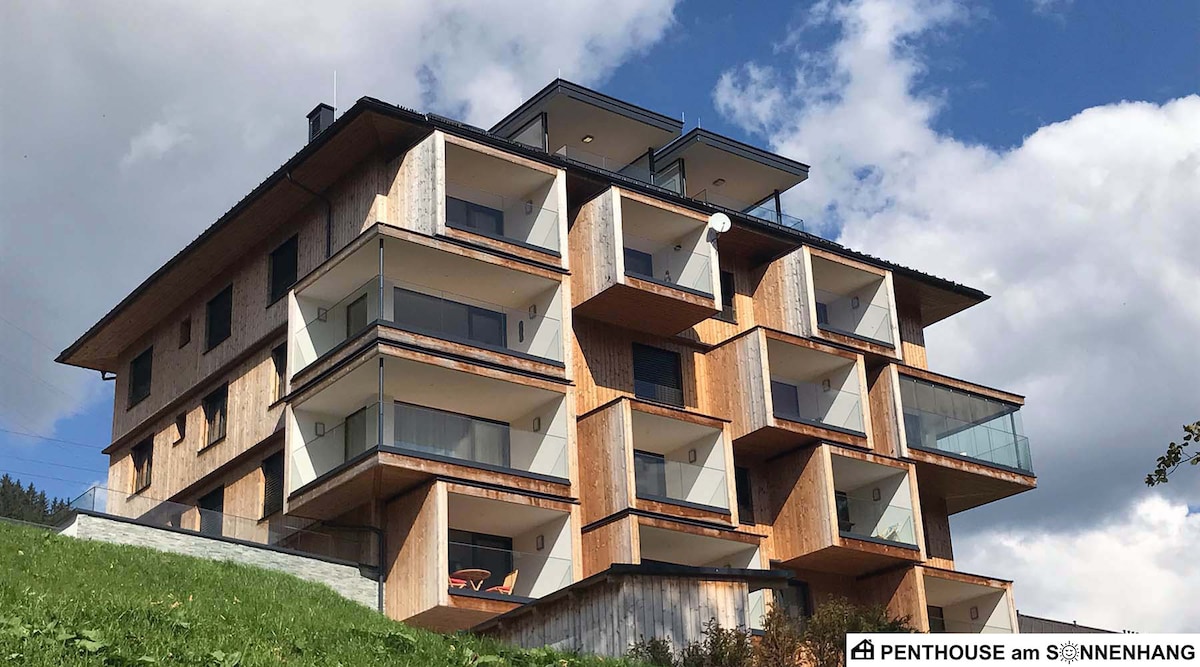 The modern building features a combination of wooden and glass elements, arranged in a staggered design. Balconies with glass railings are visible on different levels, overlooking a green landscape. The roof has a slight overhang, with a serene sky in the background.