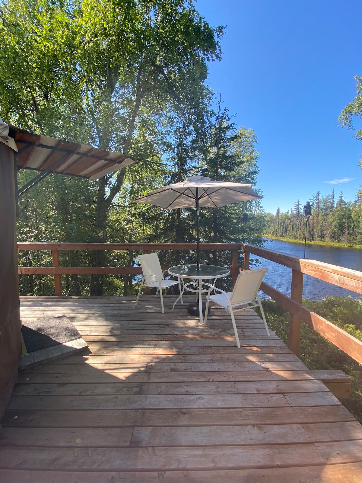 A wooden deck overlooks a serene lake, featuring a round patio table with four chairs and a large umbrella for shade. Lush trees frame the scene, providing a sense of privacy and connection to nature.