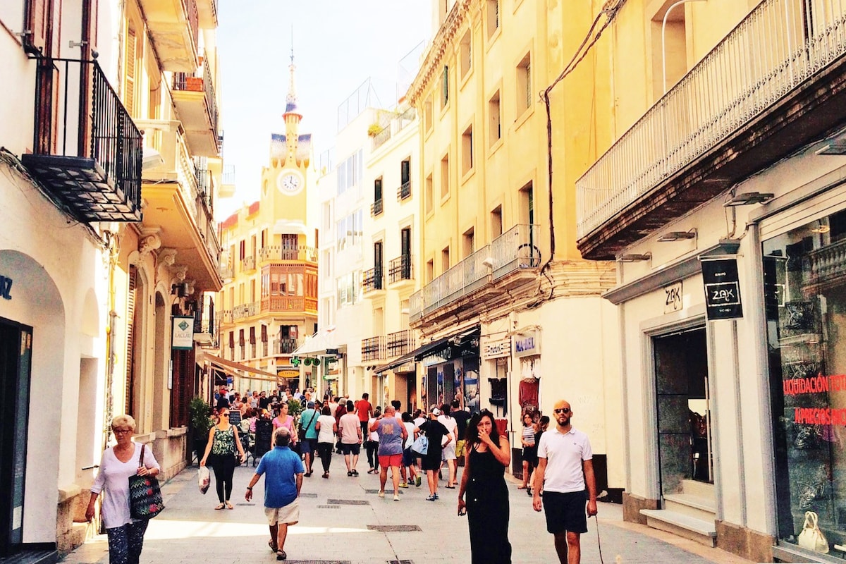 A lively pedestrian street is filled with people strolling between shops and cafes. Historic buildings line both sides, showcasing a mix of architectural styles. In the background, a clock tower stands prominently, adding character to the vibrant atmosphere of the area.