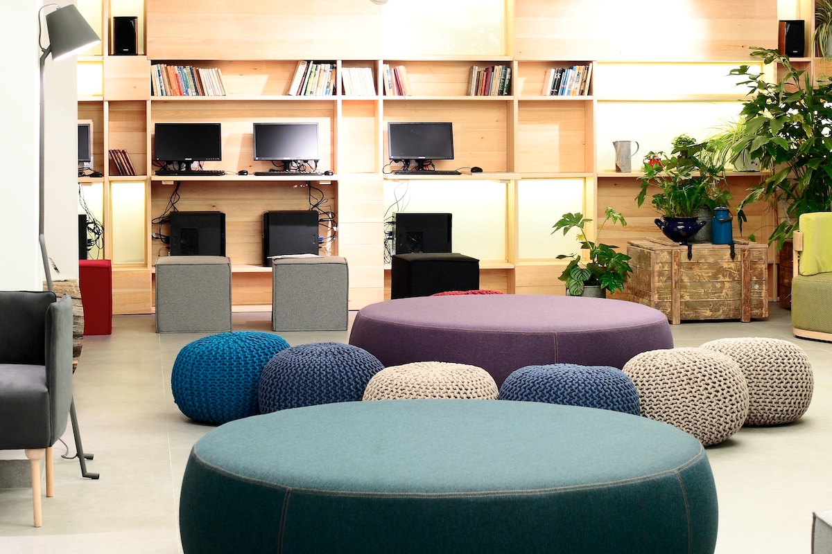 A communal area features colorful poufs in varied sizes, arranged around a round upholstered ottoman. In the background, a spacious bookshelf is lined with books, while several workstations are equipped with desktop computers. Lush greenery adds a vibrant touch to the modern setting.