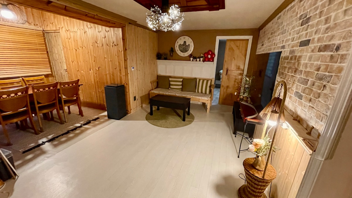A spacious living area features wooden accent walls and natural light. A low coffee table is centered on a light-colored floor, surrounded by a cozy sofa and a small reading chair. A dining area with wooden chairs is visible in the background, highlighted by a unique chandelier.