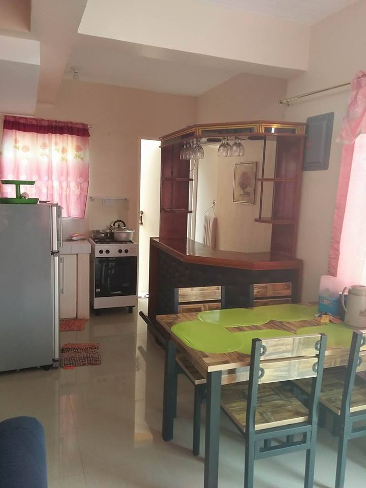 An open kitchen area features a modern stove and a refrigerator, complemented by a wooden counter bar. A simple dining table with chairs is positioned nearby, with bright curtains framing the window, allowing natural light to fill the space.