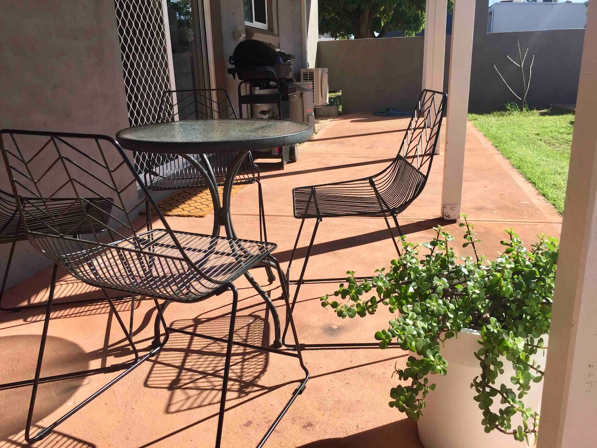 An outdoor patio area is presented, featuring a round glass table accompanied by two black wire chairs. A potted green plant adds a touch of nature, while a barbecue grill is visible in the background. Sunlight casts soft shadows across the space.