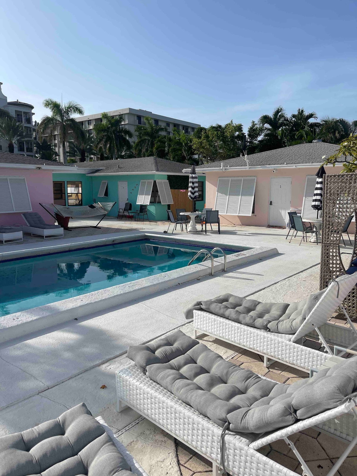 The pool area is surrounded by colorful units, featuring lounge chairs and tables arranged around the water. Palm trees provide a backdrop, and the sun shines brightly over the serene setting. Safety railings are positioned around the pool for added security.