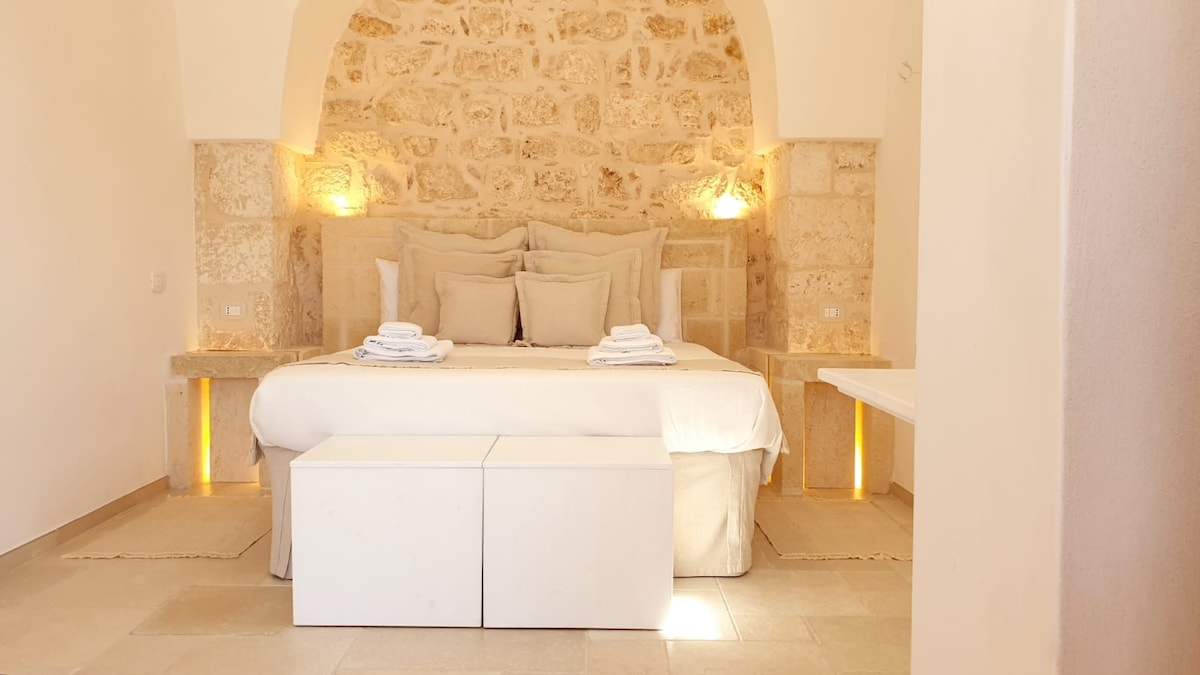 A spacious sleeping area features a large bed adorned with multiple pillows and a light blanket. Stone walls provide a natural backdrop, while soft lighting is integrated into the stone recesses. A minimalist white bench is positioned at the foot of the bed.