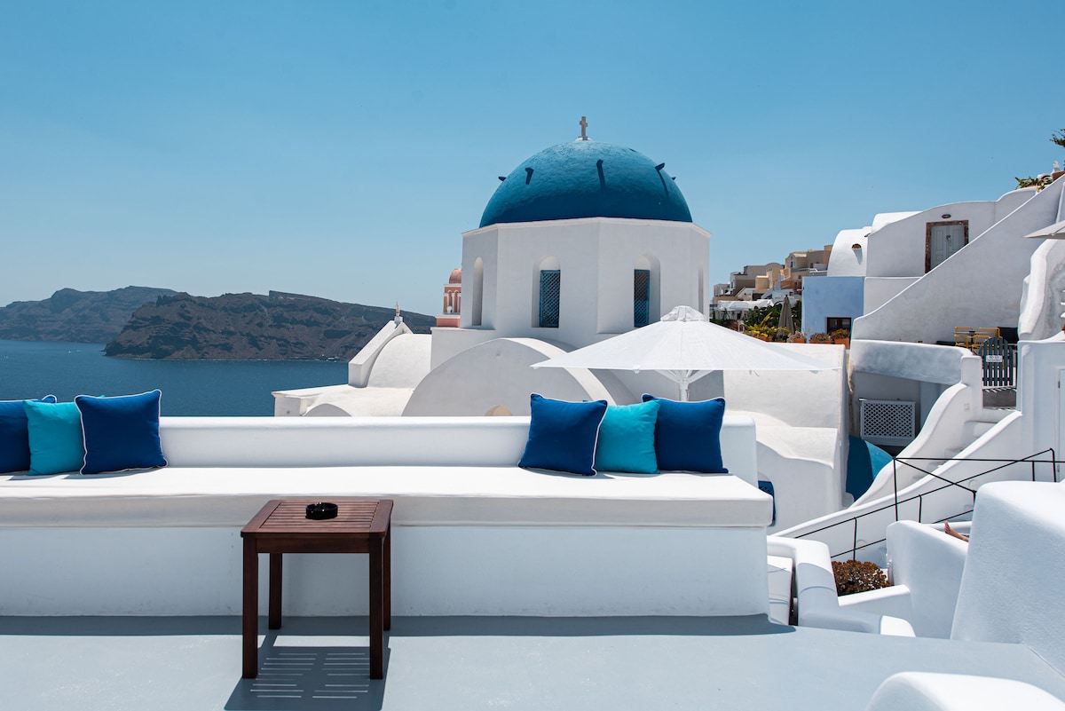 A private balcony offers a seating area with white cushions against a backdrop of the iconic blue dome and expansive sea views. A wooden table sits in front, while the surrounding white architecture enhances the serene atmosphere of the space.