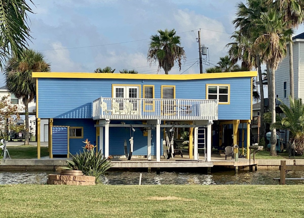 A two-story blue house with yellow trim is positioned alongside a canal. A spacious deck with white railings extends from the front, providing access to outdoor seating. Lush palm trees and other greenery are present nearby, contributing to the home's coastal ambiance.