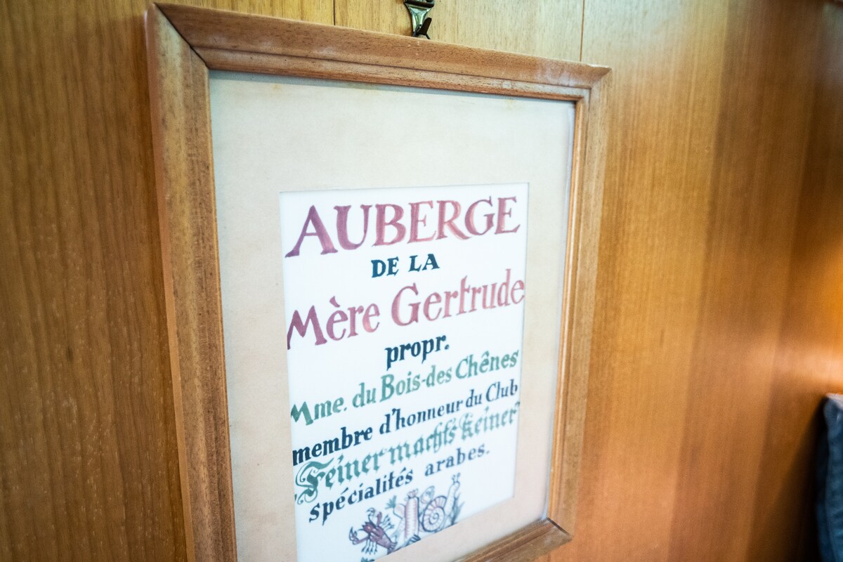 A framed poster is displayed on a wooden wall, featuring the text 'AUBERGE DE LA Mère Gertrude.' The poster contains colorful illustrations, highlighting a rustic charm. A subtle light illuminates the frame, enhancing its visibility while the surrounding wood adds a warm tone to the atmosphere.