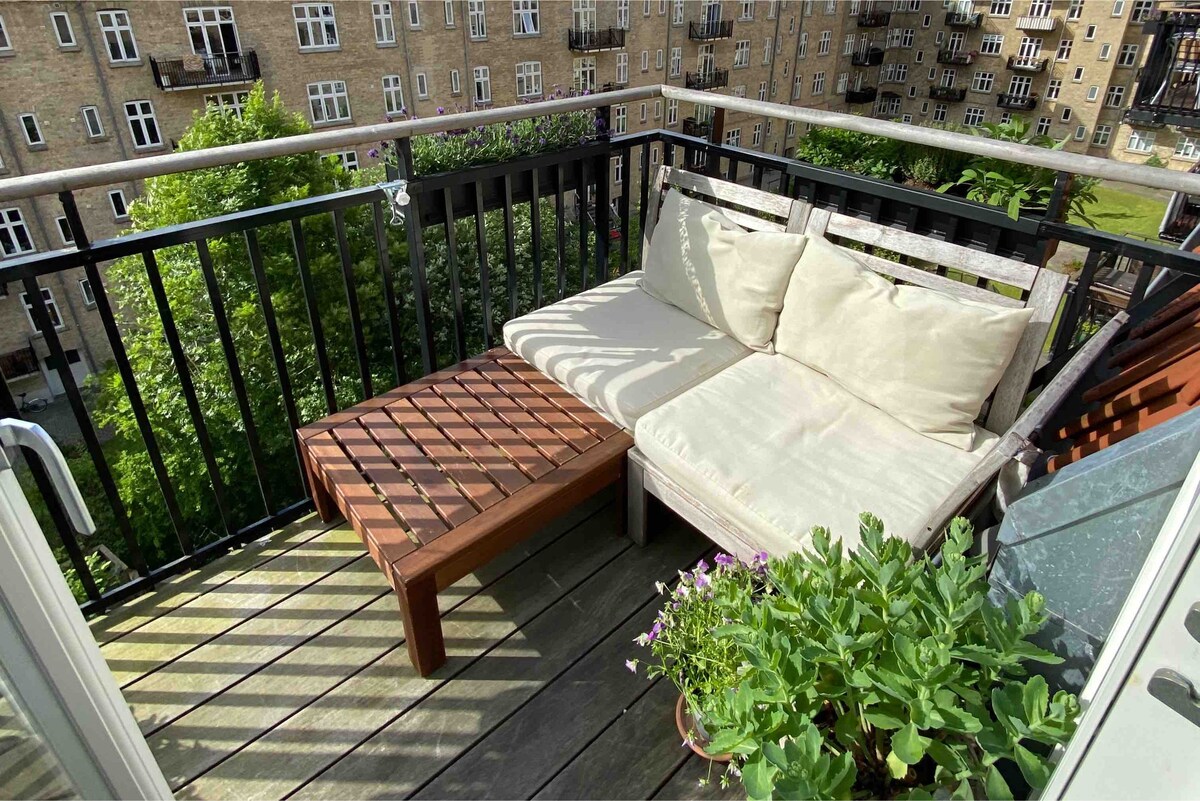 A cozy outdoor seating area is set on a wooden balcony, featuring a cushioned lounge chair and a small wooden table. Green plants are positioned on the floor, enhancing the space with natural elements. The view includes adjacent buildings and lush greenery.