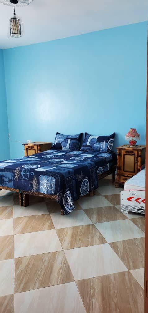 A sleeping area features a blue-themed bedspread on a spacious bed, complemented by matching pillows. Wooden nightstands are positioned on either side, with a lamp on one. The walls are painted a calming blue, and the floor showcases a patterned tile design.