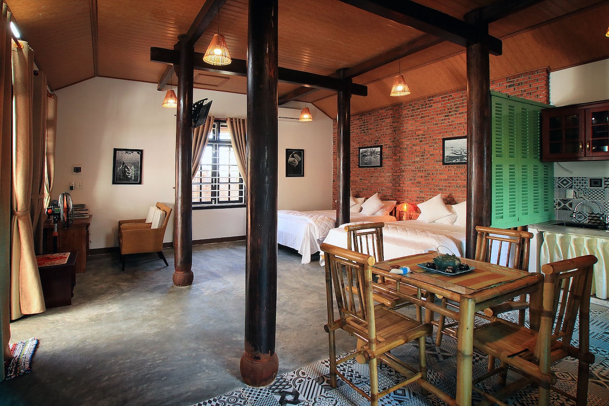 The interior of the wooden house features spacious living areas with two Queen-sized beds positioned beside large windows. Natural light filters through soft curtains, while bamboo furniture lends a traditional touch. The kitchen area can be seen in the background, fitted with modern appliances.