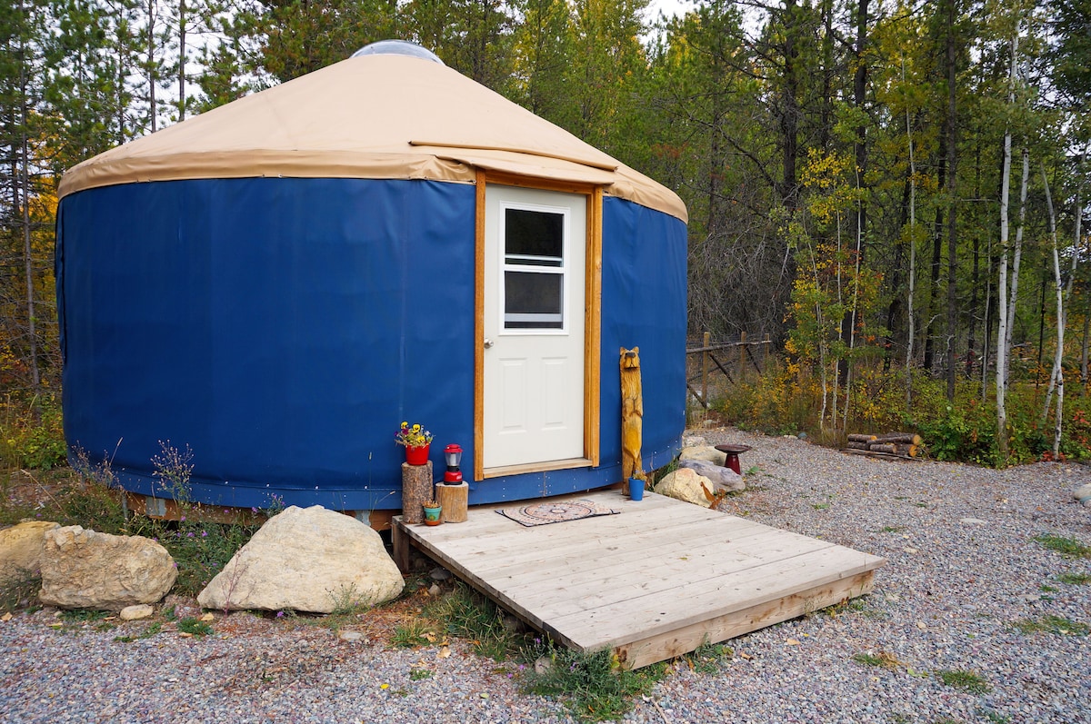 A circular yurt stands surrounded by trees, featuring a blue exterior with a tan roof. A wooden deck leads to a white door, while natural stone and pebbles border the entrance. The outdoor area is adorned with planters and a welcome mat.