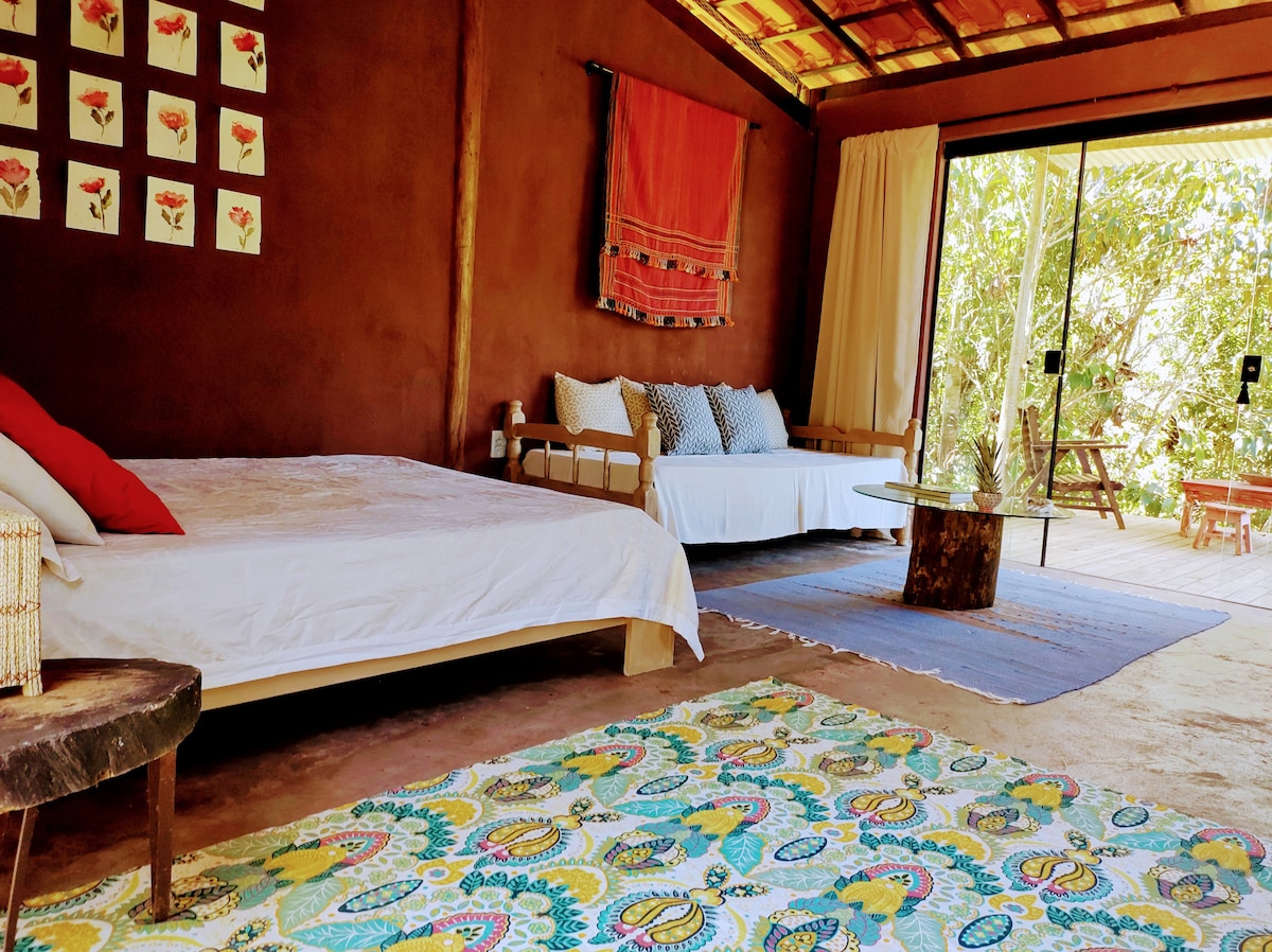 A spacious room features a comfortable bed with neutral bedding and decorative pillows. A cozy seating area includes a sofa and a wooden table. Bright artwork adorns the walls, while a colorful rug adds vibrancy to the natural tones of the space. Large glass doors open to lush greenery.
