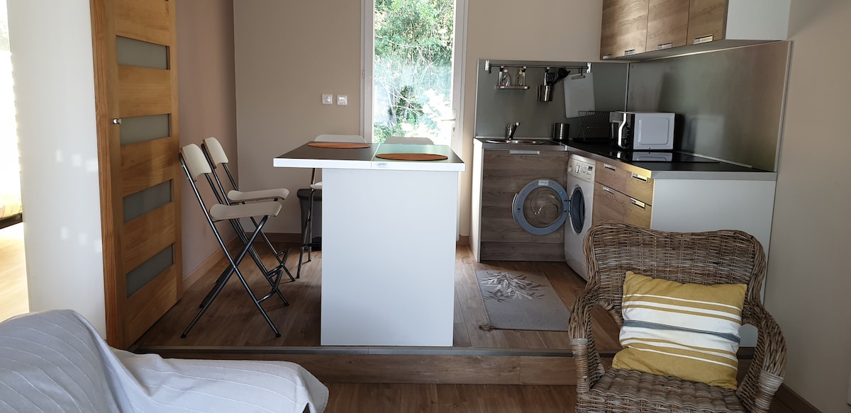 A modern kitchenette is equipped with stainless steel appliances and a washing machine. Two bar stools are positioned at the island, while a cozy armchair with decorative pillows is nearby. Natural light enters the space through a window, enhancing the warm wood tones of the flooring.