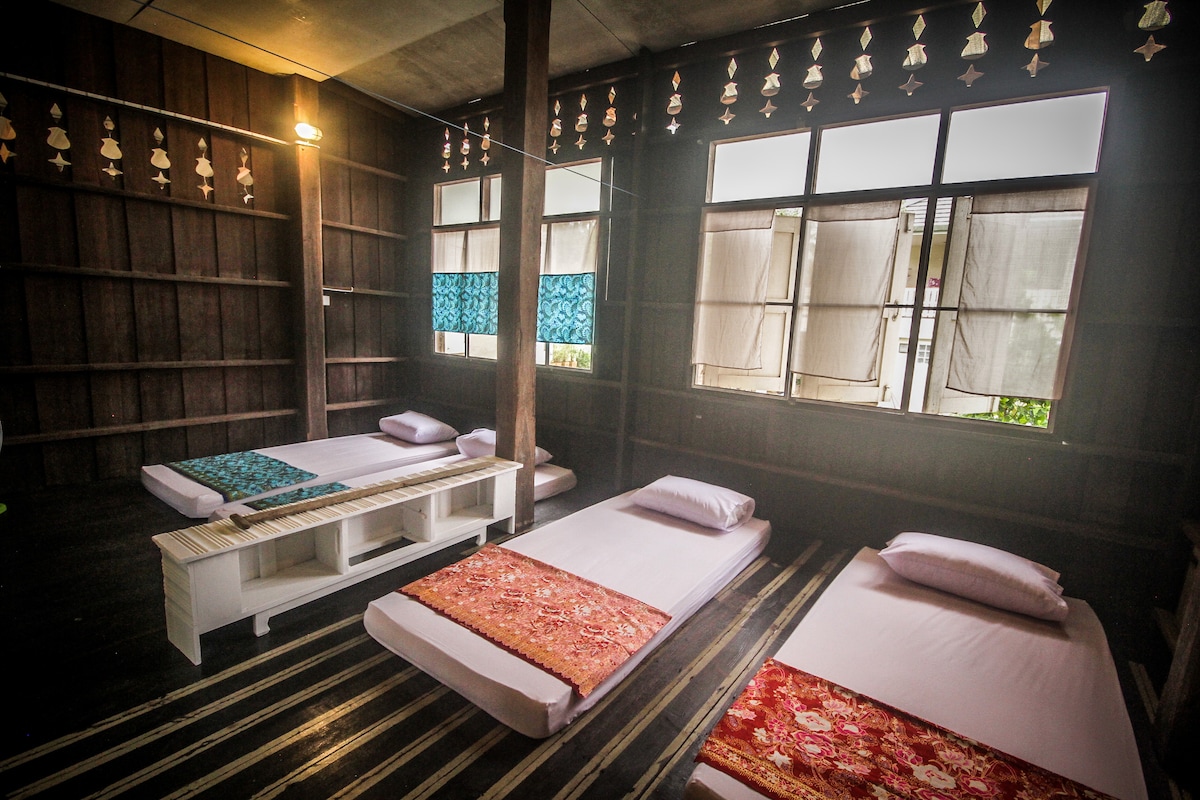 A spacious room features four set sleeping areas arranged with comfortable mattresses on a wooden floor. Colorful fabric decorates each mattress, and natural light enters through large windows adorned with light-curtain panels. Shelves line one wall, enhancing the room's open feel.