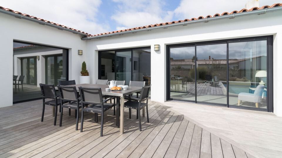 A spacious wooden deck is featured, offering an outdoor dining area with a large table and several black chairs. Large glass doors provide views into the interior of the house, while glimpses of a swimming pool and garden are visible in the background.