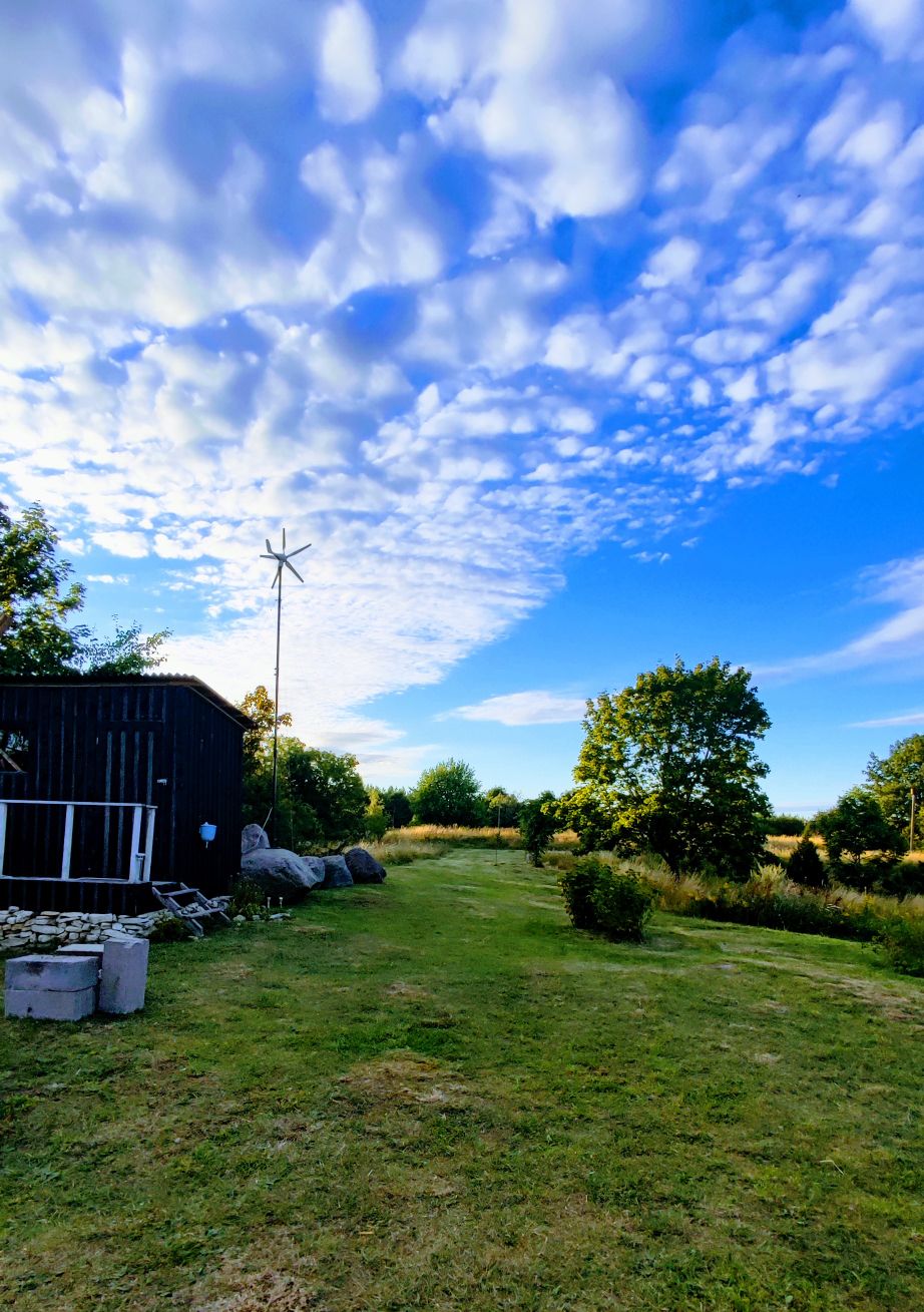 A serene outdoor area features a well-maintained grassy lawn surrounded by trees and shrubs. A small structure is visible on the left, with a wind turbine standing tall against the bright blue sky filled with scattered clouds.