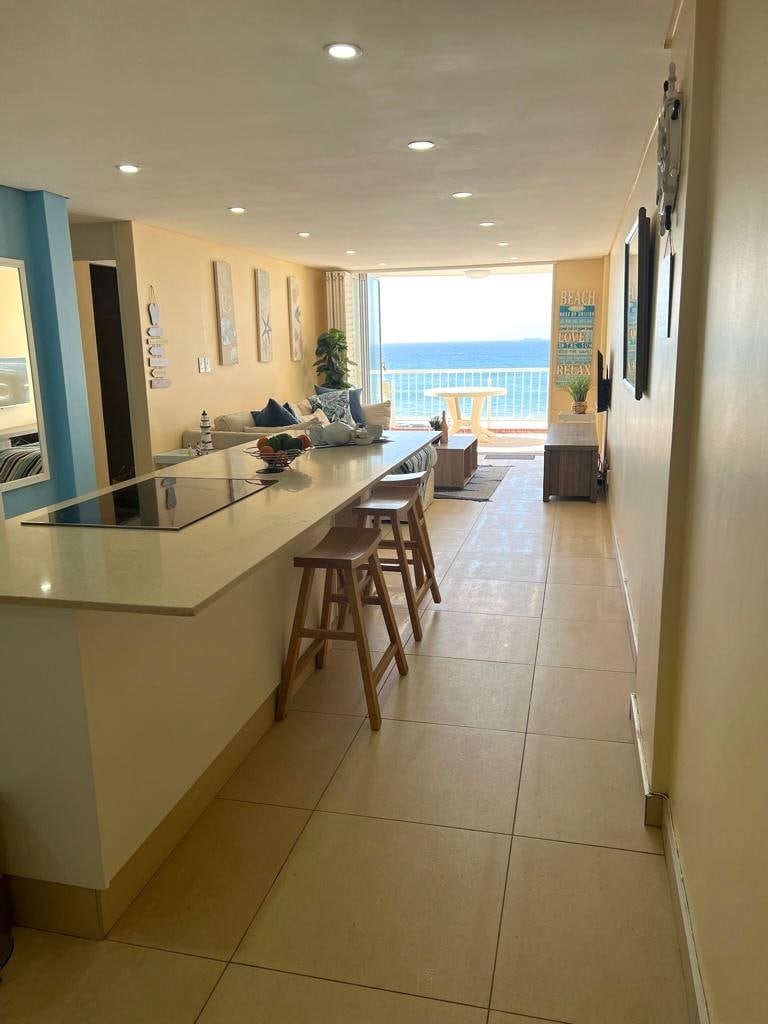 An open-plan living area is visible, featuring a kitchen with a breakfast bar and wooden stools. Natural light floods the space through large sliding doors that offer views of the ocean. A lounge area with comfortable seating is present, creating an inviting atmosphere.