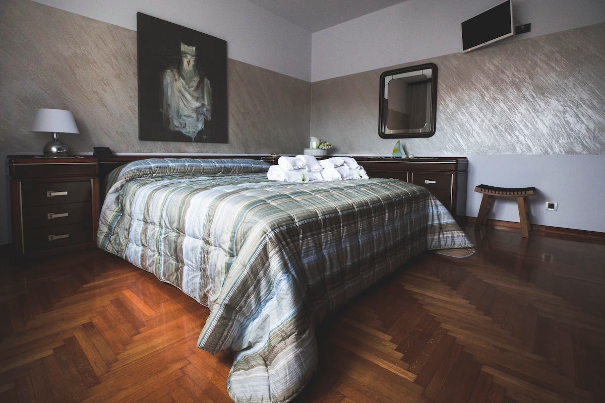 A spacious bedroom features a patterned bedspread and a variety of pillows neatly arranged on the large bed. A simple wooden stool is placed nearby. Natural light creates a warm ambiance, while a large artwork and wall mirror add to the room's decor.