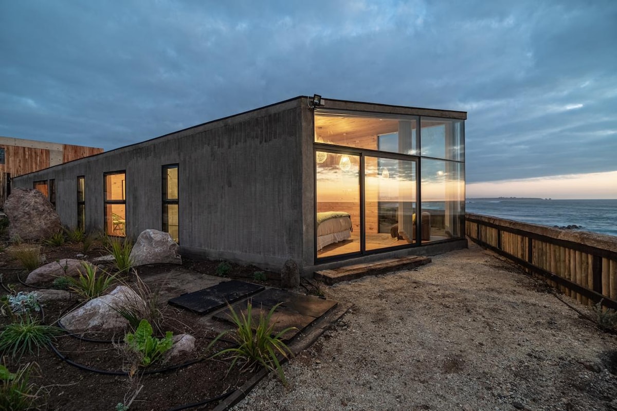 A modern, single-story home features large glass windows that offer views of the ocean. The exterior is designed with a combination of concrete and natural materials, surrounded by smooth stones and low vegetation. The structure's clean lines create a harmonious integration with the landscape.