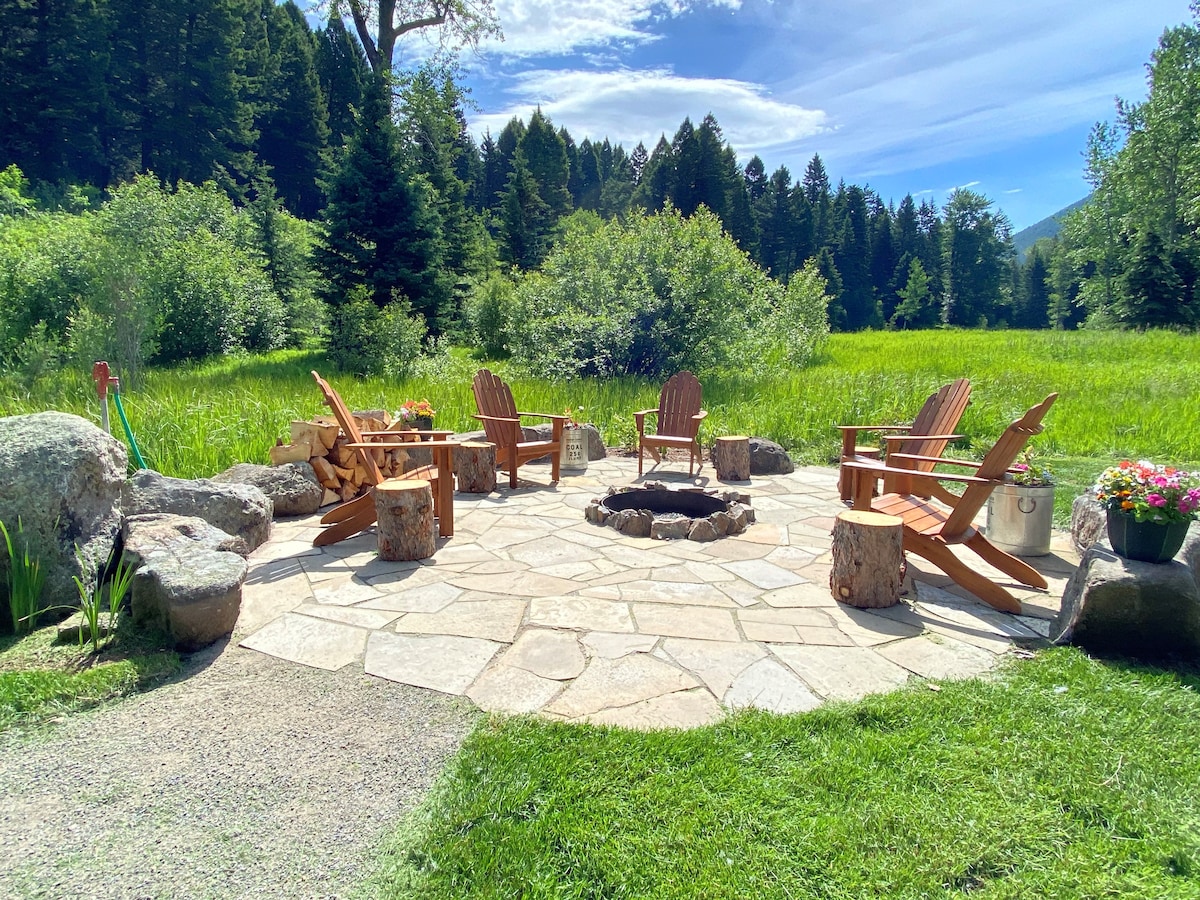A stone patio features a circular arrangement of wooden Adirondack chairs surrounding a central fire pit. Natural greenery and trees form a backdrop, enhancing the outdoor setting. Stone and wooden accents contribute to the rustic charm of the area.