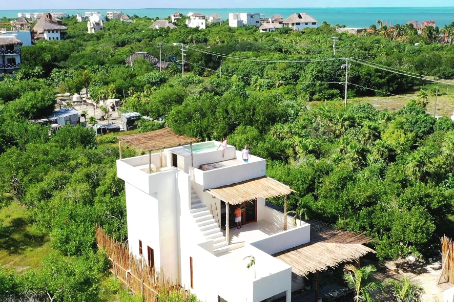 The villa is positioned amidst lush green vegetation, offering a private rooftop terrace with a pool. The layout features multiple levels, showcasing a modern design that blends seamlessly into the surrounding landscape. Ocean views are visible in the distance, enhancing the tranquil atmosphere.