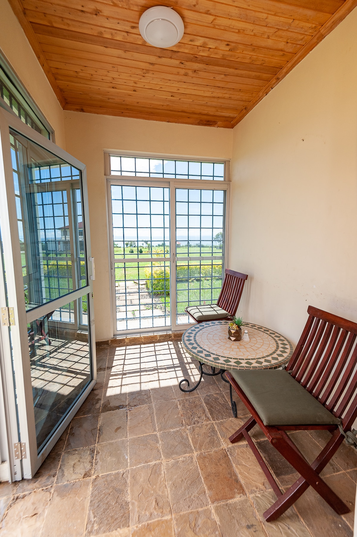 A serene sitting area features two wooden chairs and a round table with intricate mosaic design. Large glass doors open to a view of the outdoors, allowing natural light to illuminate the space.