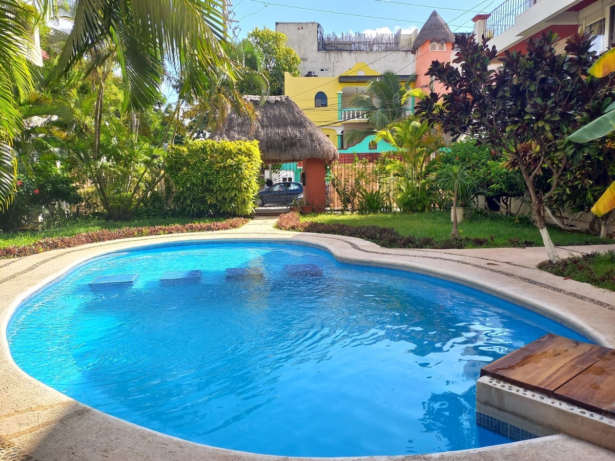 A tranquil swimming pool is surrounded by lush greenery and vibrant landscaping. The pool features a gentle curve and is enhanced by sunlight reflecting off its clear blue water. Colorful buildings can be glimpsed in the background.