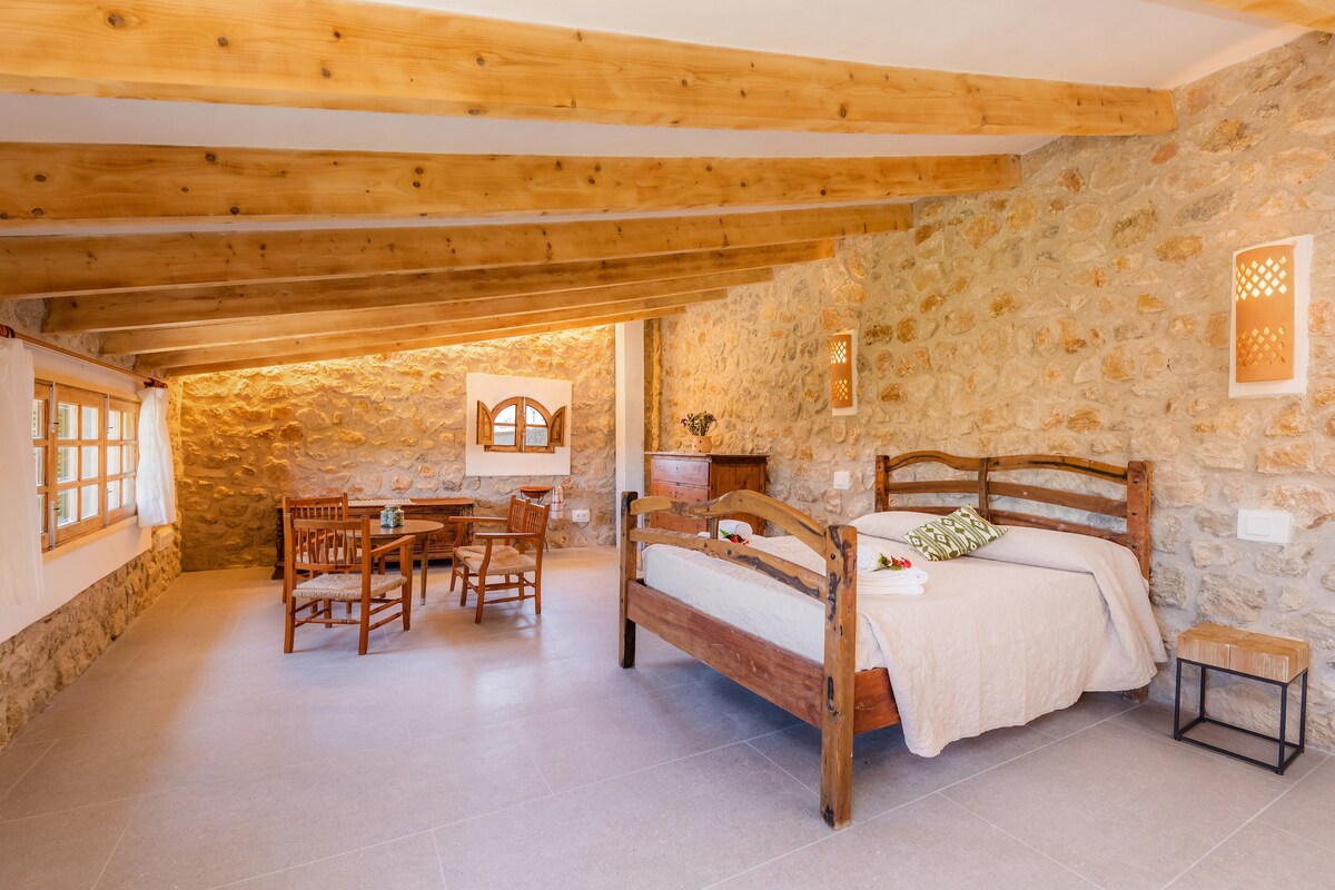 A spacious bedroom features a wooden bed frame adorned with neutral linens. Natural stone walls complement the rustic vibe. A small wooden table and chairs provide a cozy dining space, while soft lighting enhances the room's inviting ambiance.