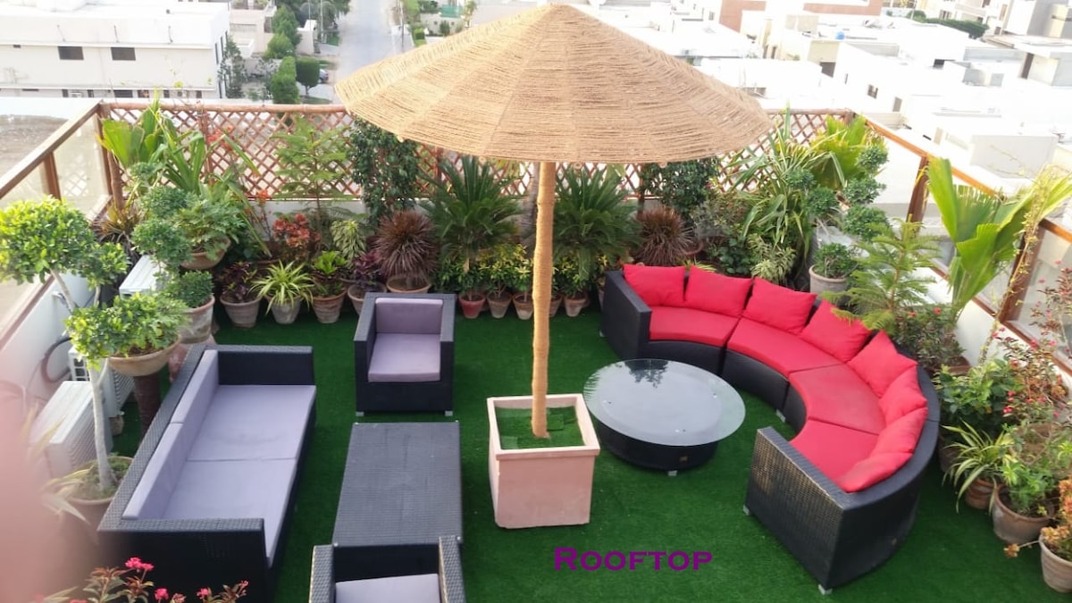 An inviting rooftop terrace features a shaded straw umbrella above a round glass table. Surrounding seating consists of a circular arrangement of red cushioned sofas and additional low-backed chairs. Lush greenery in varied planters enhances the outdoor space, offering a comfortable setting for relaxation.