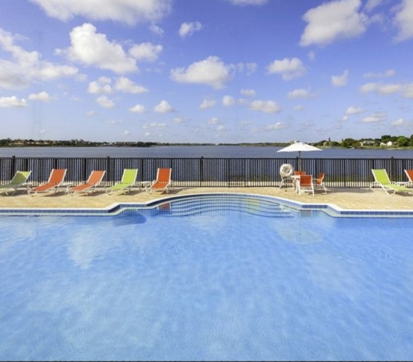 A refreshing swimming pool is visible in the foreground, surrounded by lounge chairs in vibrant colors. Beyond the pool, a scenic view of a large body of water is framed by a black railing, with fluffy clouds adding texture to the blue sky.